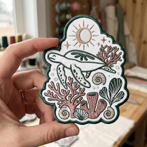 May include: An embroidered patch featuring a white sea turtle swimming among pink coral, sea sponges, and shells. Above the turtle, a golden sun and moon design is surrounded by small stars. The entire patch is outlined in a dark green border.