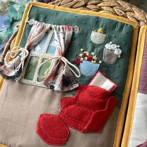 Puede incluir: Un libro de juego de fieltro en forma de casa con un sillón rojo y un reposapiés, una ventana con cortinas y flores bordadas en macetas.