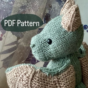 May include: A knitted green and brown dragon toy with a PDF pattern. The dragon has a long tail and wings. The toy is sitting on a woven basket.