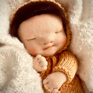 Könnte beinhalten: Eine handgefertigte Stoffpuppe mit braunen Haaren, die ein gestricktes gelbes Outfit trägt, schläft friedlich in einer weißen Decke.