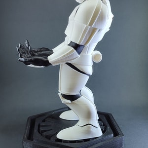 May include: A white 3D printed Star Wars Stormtrooper figurine standing on a black hexagonal base with the Star Wars logo. The figurine is posed with its arms outstretched.
