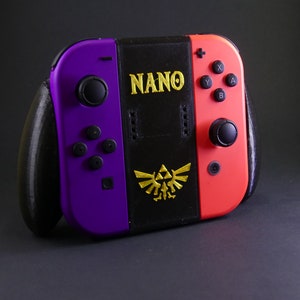 Op de afbeelding: Een paarse en rode Nintendo Switch-console met zwarte joy-cons en een zwarte frontplaat. De frontplaat heeft het woord "NANO" en een gouden Triforce-symbool.