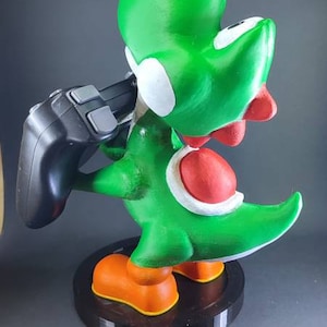 Op de afbeelding: Een groene en rode Yoshi-figuur die een zwarte videogamecontroller vasthoudt. De figuur staat op een zwarte basis.