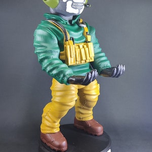 Puede incluir: Una figura verde y amarilla de un personaje del videojuego Call of Duty. El personaje lleva una máscara de calavera, una chaqueta verde y pantalones amarillos. La figura tiene las manos extendidas hacia delante. La figura está de pie sobre una base negra con la palabra "COD" escrita en ella.