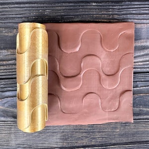 Puede incluir: Un rodillo dorado con un diseño de patrón ondulado. El rodillo se utiliza para crear un patrón ondulado en la arcilla. La arcilla es de color marrón rojizo.