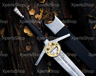 May include: A detailed image of a sword with a silver blade and a black handle. The sword features a gold-colored crossguard with green accents and a decorative emblem. The scabbard is black. The sword rests on a dark, textured surface.