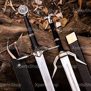 Può includere: Due spade con lame argentate e manici neri poggiano su una superficie di legno. Una spada presenta un emblema circolare sull'elsa, l'altra una testa di animale stilizzata. Entrambe hanno foderi in pelle.