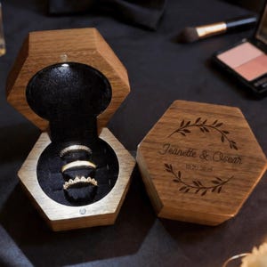 May include: A wooden, hexagonal ring box with a hinged lid, open to reveal three gold rings. The box is engraved with the names "Jeanette & Oscar" and a date. The interior is lined with black velvet.