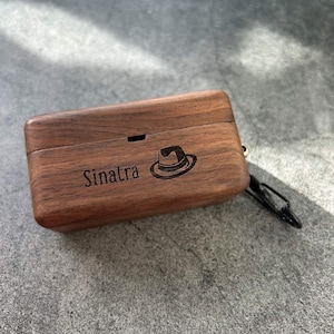 Puede incluir: Una funda rectangular de madera con el nombre "Sinatra" y un diseño de sombrero fedora grabado. La funda es de color marrón cálido y tiene un pequeño lazo negro en el lateral. El fondo es una superficie gris texturizada.