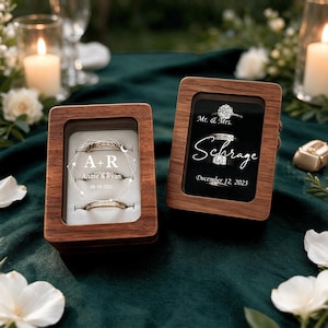 May include: Two rectangular wooden ring boxes with glass lids. The left box features white text A+R, Annie & Ryan, and 08-10-2025. The right box displays Mr. & Mrs. Schrage and December 12, 2025. Both boxes contain silver rings on white velvet inserts.