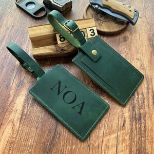 Puede incluir: Dos etiquetas de equipaje de cuero verde con una hebilla dorada. Una etiqueta tiene el nombre NOA grabado en relieve.