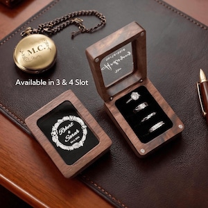 May include: Two rectangular wooden ring boxes with glass lids, one closed with a white floral wreath design and the text Robert & Sarah 04.11.2024, the other open displaying four rings in black velvet slots, alongside a gold pocket watch and a pen.