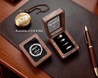 May include: Two rectangular wooden ring boxes with glass lids, one closed with a white floral wreath design and the text Robert & Sarah 04.11.2024, the other open displaying four rings in black velvet slots, alongside a gold pocket watch and a pen.