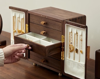 May include: A dark brown wooden jewelry box with multiple drawers and compartments. The box has a hinged door on each side, revealing hanging space for necklaces. Gold necklaces and other jewelry are visible. The drawers have leather pulls.