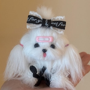 Può includere: Una piccola statuetta di cane in peluche bianco e soffice con occhi neri e lingua rosa. Presenta una molletta rosa sulla fronte, due elastici rosa sulle orecchie e un fiocco in nastro bianco e nero con la scritta "just for you" sulla testa.