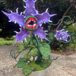 Op de afbeelding: Handgemaakte, grillige plantensculptuur met een grote, paarse, stekelige bloem en een rood-witte getande mond. De plant heeft groene bladeren, wijnstokken en staat in een zilveren metalen emmer. Een uniek decoratief item.