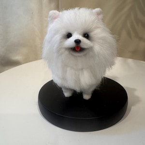 Può includere: Statuetta di un cane Pomerania bianco con occhi neri e lingua rossa, in piedi su una base circolare nera. Il cane ha un folto pelo di peluche, su sfondo neutro.