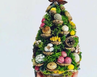 May include: A miniature Christmas tree crafted from moss, pine cones, and other natural materials. The tree is adorned with small white, pink, and yellow flowers, and a star-shaped decoration at the top.