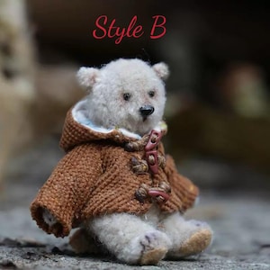 Pu&ograve; includere: Un orsacchiotto bianco che indossa un cappotto di maglia marrone con cappuccio e bottoni marroni. L'orso &egrave; seduto su una superficie strutturata. Il testo "Style B" &egrave; scritto in rosso sopra l'orso.