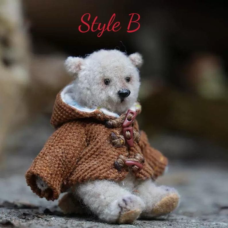 Pu&ograve; includere: Un orsacchiotto bianco che indossa un cappotto di maglia marrone con cappuccio e bottoni marroni. L'orso &egrave; seduto su una superficie strutturata. Il testo "Style B" &egrave; scritto in rosso sopra l'orso.