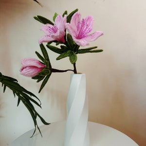 May include: A bouquet of pink azalea flowers with purple accents in a white, twisted vase. The flowers are arranged in a simple, elegant style.