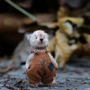 May include: A small, white teddy bear wearing brown dungarees with four buttons. The bear has a brown nose and black eyes. The bear is standing on a grey surface.