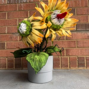 May include: A decorative floral arrangement featuring two large, yellow sunflowers with orange tips and green centers. The flowers are in a gray cylindrical pot with green leaves.