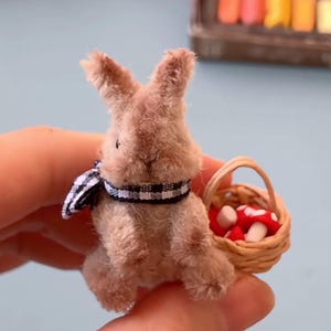 May include: A miniature brown bunny figurine wearing a black and white checkered scarf. The bunny holds a small woven basket filled with red and white mushroom figurines. The bunny is made of a soft, plush material.