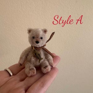 Pu&ograve; includere: Un piccolo orsacchiotto di peluche beige con un cinturino in pelle marrone intorno al collo. Il cinturino ha un ciondolo a forma di pesce dorato. L'orsacchiotto &egrave; seduto su una mano, e il testo "Style A" &egrave; scritto in rosso sopra l'orsacchiotto.