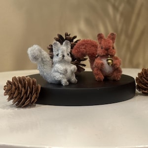 Puede incluir: Dos pequeñas figuras de ardillas peludas, una gris claro y otra marrón rojizo, están colocadas sobre una base circular negra. La ardilla marrón rojizo lleva un pequeño dije de bellota dorado en el cuello. Hay tres piñas marrones.