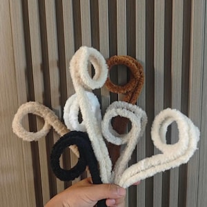 May include: A collection of fluffy, looped craft supplies in various colors, including white, black, brown, and beige. The soft, textured material is held against a neutral, vertical-striped background.