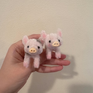 Puede incluir: Dos pequeñas figuras de cerdos rosas de peluche con ojos negros y hocicos color canela. Los cerdos tienen orejas y patas rosas. Las figuras se sostienen en una mano sobre un fondo neutro.