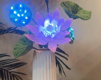 May include: A white ceramic vase with a textured surface holds a bouquet of pink and white artificial lotus flowers with blue LED lights. The flowers are surrounded by green leaves.