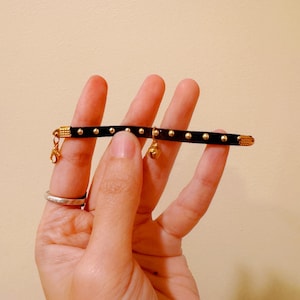 Op de afbeelding: Een zwarte leren armband met gouden accenten en een klein gouden belletje.