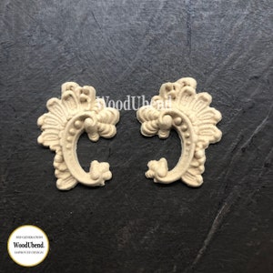 May include: Two ornate, cream-colored wooden embellishments with a swirling, floral design. The embellishments are shaped like a stylized letter C.