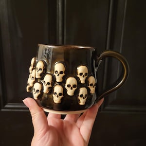 May include: A dark brown ceramic mug featuring a repeating pattern of small, raised, off-white skull motifs encircling the exterior. The mug has a glossy finish, a curved handle on the right side, and a plain, unglazed light-coloured base rim.
