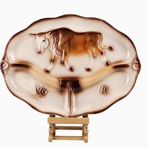 May include: An oval ceramic serving platter featuring a raised brown bull figure in the center. The platter has a cream-colored background with brown airbrushed edges and a rope-like border. The letters "LO" and "BO" are embossed on the lower section.