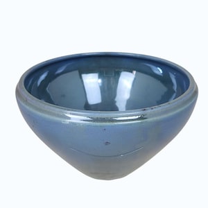 May include: A blue ceramic bowl with a rounded rim and a smooth, glossy interior. The bowl has a slightly flared shape and a matte exterior. The color is a muted blue-gray, with subtle variations in tone. The bowl is likely used for serving or display.