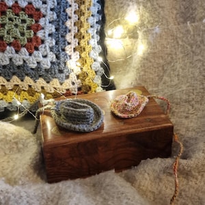 May include: Two small crocheted cowboy hats, one gray and one pink, are sitting on a wooden box. The box is on a white crocheted blanket.