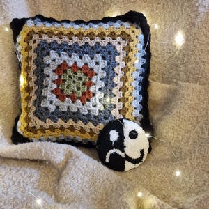 Puede incluir: Una almohada tejida a crochet con un patrón de cuadrado de la abuela colorido y un símbolo yin yang en blanco y negro con una cara triste en un lado y una cara feliz en el otro. La almohada está sobre una manta beige con luces blancas.