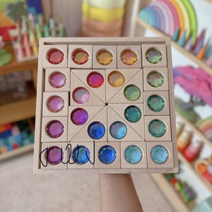 May include: A square wooden tray containing a collection of light-coloured wooden blocks with circular, faceted, iridescent acrylic inserts in various colours including red, orange, yellow, green, blue, and purple, arranged in a geometric pattern.