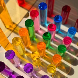 Könnte beinhalten: Eine lebendige Anordnung farbiger, zylindrischer Objekte auf einer weißen, geriffelten Oberfläche. Die Objekte sind in Rot-, Blau-, Grün-, Gelb-, Orange- und Lilatönen gehalten. Das Licht erzeugt Schatten, die Farben und Formen hervorheben.