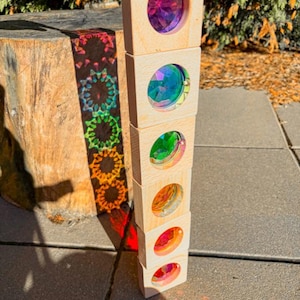 Puede incluir: Un juguete de madera con seis bloques, cada uno con una lente iridiscente de colores. Los bloques están apilados verticalmente, proyectando un patrón de arcoíris sobre una superficie cercana. El juguete está diseñado para el juego educativo.