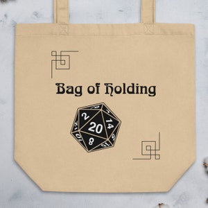 May include: A beige canvas tote bag with black handles and the text "Bag of Holding" printed on the front. A black and white 20-sided die is centred below the text.