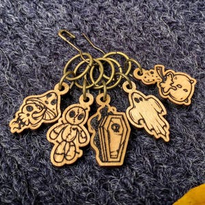 Op de afbeelding: Een verzameling houten steekmarkeerders met Halloween-thema's. De markeringen tonen een kat, een voodoopop, een kist, een spook en een pompoen. De markeringen zijn bevestigd aan bronskleurige ringen en een veiligheidsspeldsluiting.