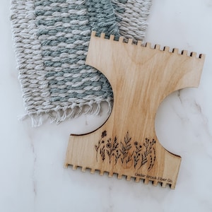 Pode incluir: Um tear de madeira com um design floral e o texto "Cedar Brook Fiber Co.". O tear é usado para criar uma tapeçaria tecida com um padrão azul e branco.