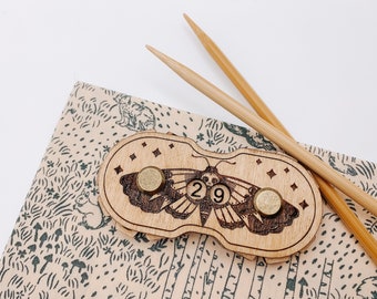 May include: Wooden moth-shaped stitch marker with brass accents and the number 29. Two bamboo knitting needles rest beside the marker on a beige fabric with a forest pattern. The marker has a skull design.