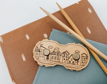 May include: Wooden stitch counter with a city design and two brass knobs. The counter displays the numbers "63". The counter is on a brown fabric with white stripes and a blue fabric.