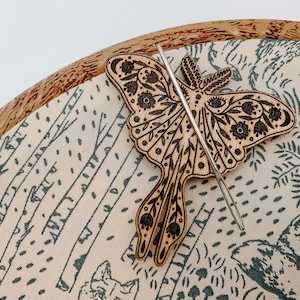 Könnte beinhalten: Ein hölzerner Schmetterlings-Stickrahmen-Nadelhalter mit floralem Design. Der Nadelhalter liegt auf einem cremefarbenen Stoff mit grünem Waldmuster.