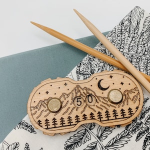 以下が含まれることがあります： 木製の山をモチーフにしたカウンター。金属製のリングが2つと、数字の「50」が刻まれています。カウンターは、黒いシダ模様の白い布の上に置かれています。